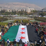 TOPSHOT - People hold a large Syrian opposition flag at Umayyad Square in Damascus on December 9, 2024. Syrians flocked to the main square of the capital city Damascus on December 9 to mark what many regard as a long-awaited new dawn after the fall of president Bashar al-Assad. Assad fled to Russia the day before after a lightning offensive spearheaded by Islamist rebels ousted him from power, opening a new chapter in Syria's history after five decades of rule by his clan. (Photo by Omar HAJ KADOUR / AFP) (Photo by OMAR HAJ KADOUR/AFP via Getty Images)